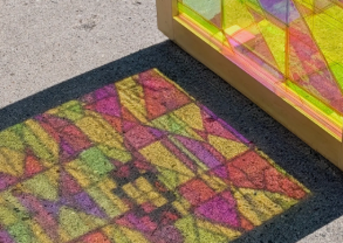 Sunlight shining through a colorful geometric stained-glass-style panel casts a vivid pattern of pink, yellow, green, and purple shapes onto a dark surface. The shadow creates a mosaic-like design of overlapping triangles and rectangles on the pavement beside the panel.