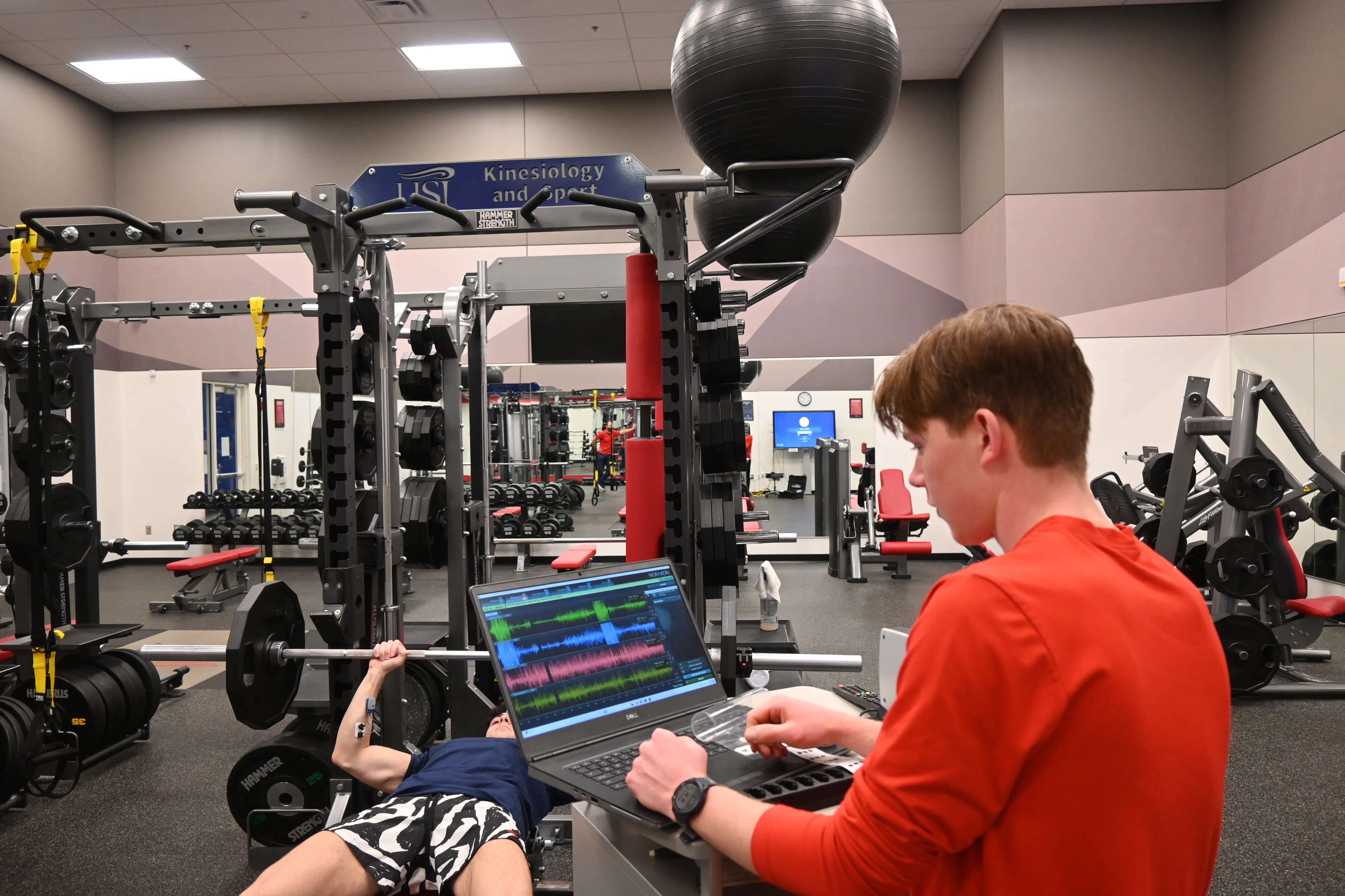 Kinesiology and sport student works on a laptop while observing someone benchpressing