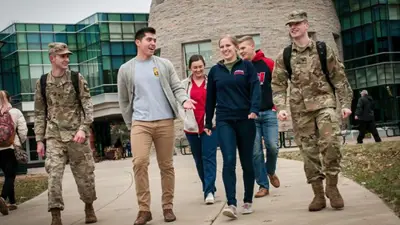 A group of students walk in front of the University Center, some of them in street clothes and some of them in military uniform