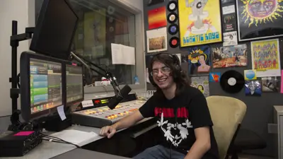 Student DJ smiles from behind the soundboard at the radio studio, which is covered in music posters