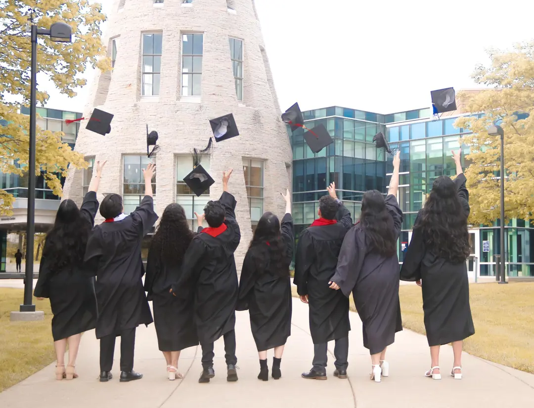 International students celebrate graduation by tossing their caps in the air outside a USI academic building. Wearing commencement gowns, the graduates stand on a campus walkway with the building’s stone tower, glass windows, and landscaped grounds visible in the background.