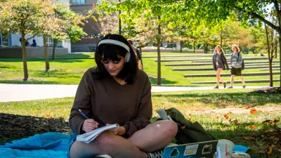 Student sits on a blanked on the Quad writing in a notebook and wearing headphones