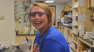 Female medical sciences student smiles while conducting a lab experiment with pipettes, latex gloves, and safety goggles
