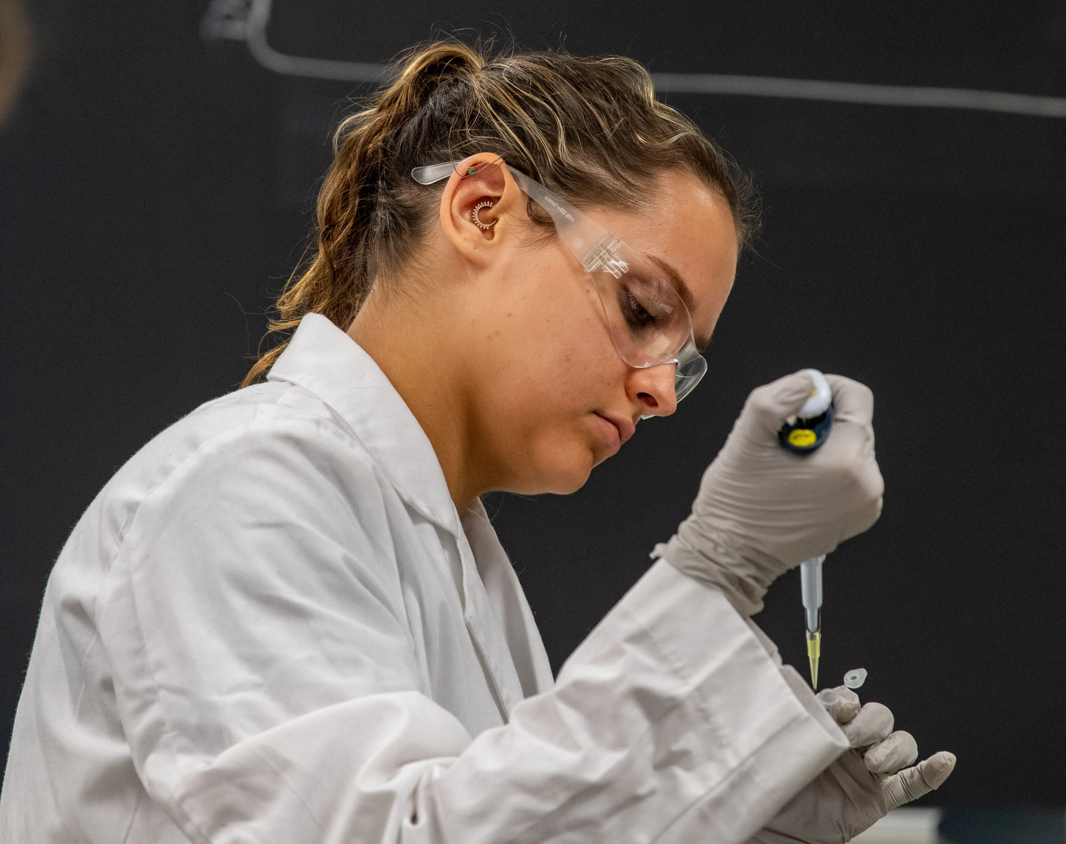 Student wearing a lab coat and safety glasses uses a pipette to add liquid to a test vial in the genetics lab