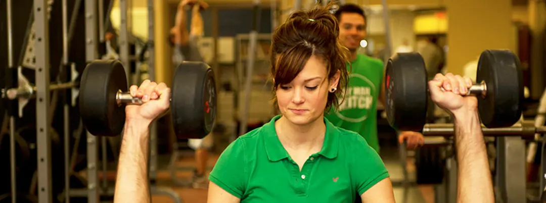 Female kinesiology student supports a weight lifter in the gym