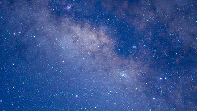 Wide view of a star-filled night sky with the Milky Way visible as a dense, cloudy band of stars stretching across the image in shades of blue and purple.