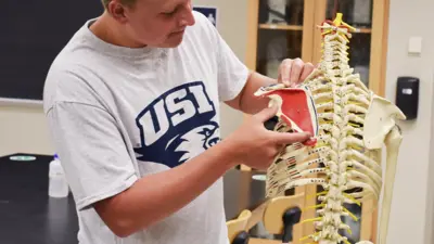 Student in a USI t-shirt holds a scapula up to its location on a model human skeleton