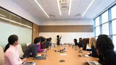 An instructor writes on the whiteboard while a group of professionals seated around a conference table observe