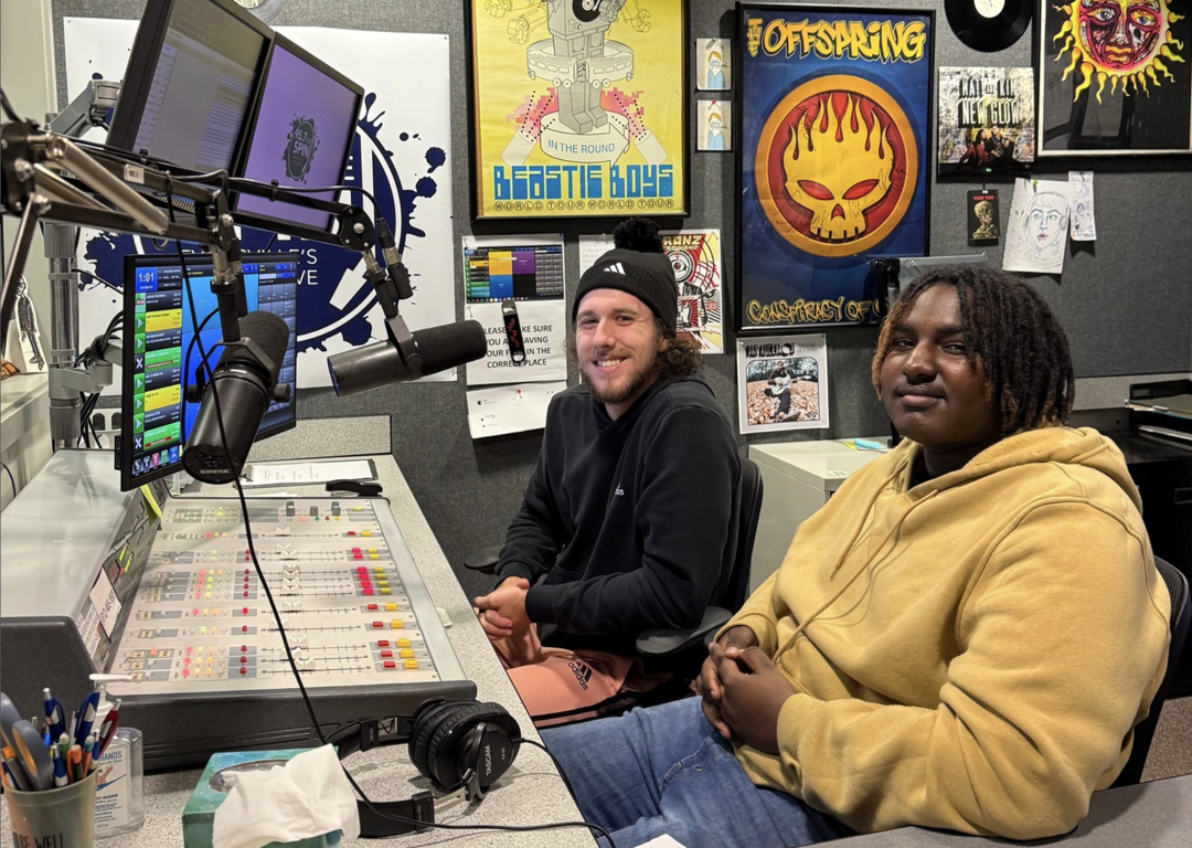 Two male students smile at the camera from behind the soundboard in the USI campus radio studio