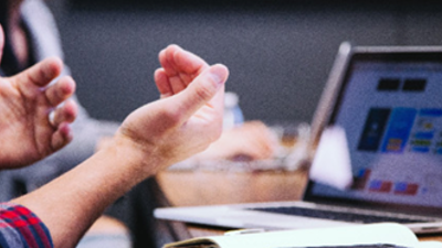 A close-up of someone's hands making gestures with a laptop screen in the background