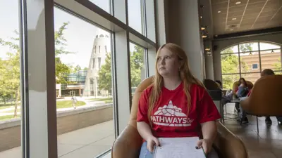 Student in a Pathways to USI t-shirt sits in the Rice Library lounge by a window with the University Center Tower visible in the background