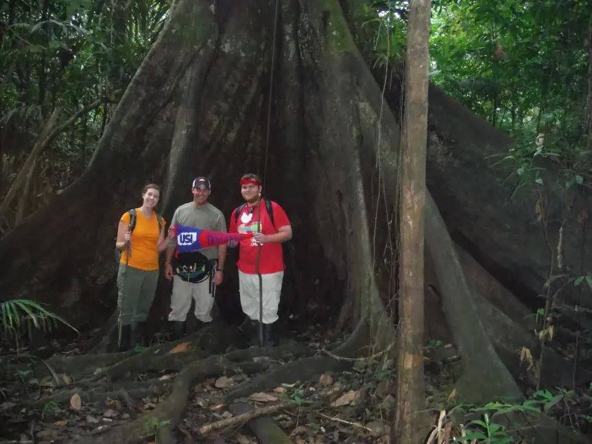 Anthropology faculty and students pose with a USI banner at the base of a very large tree in the rainforest