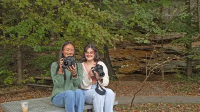 Students with cameras sit on an outdoor picnic table on campus