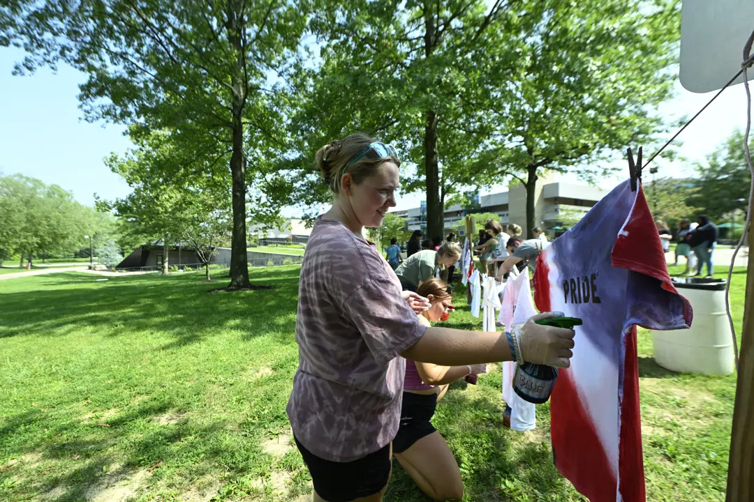 Students spray colorful dye onto "Pride" t-shirts hanging from an outdoor clothesline
