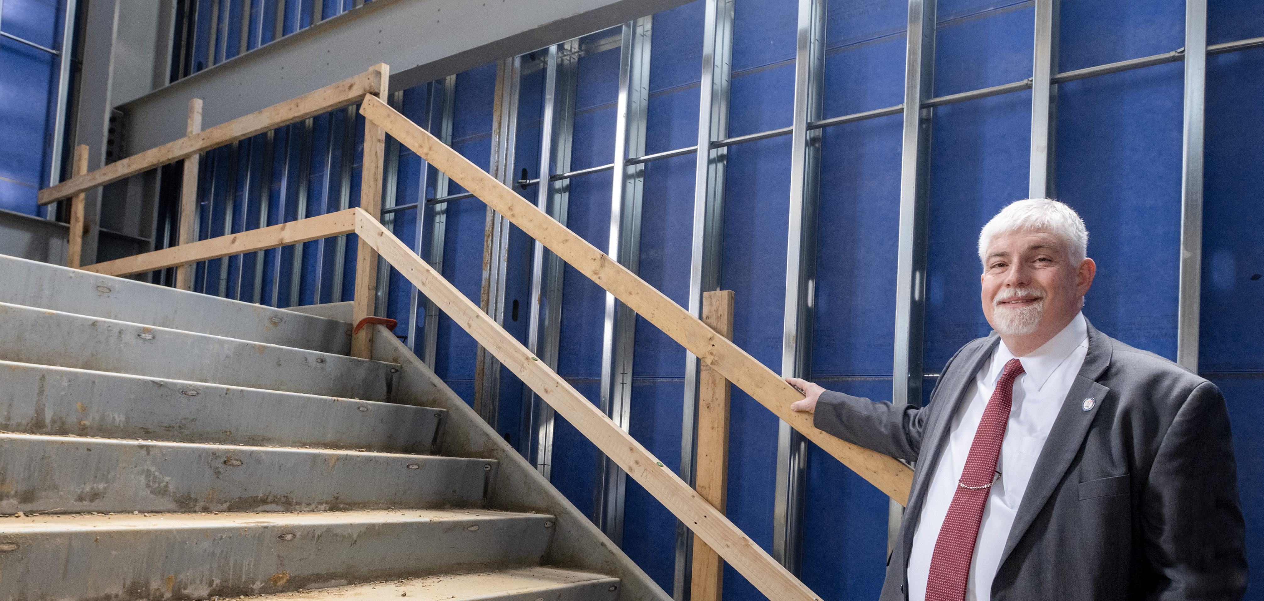 President Bridges wears a suit tie while standing on an under-construction staircase in the Health Professions Center renovation site.